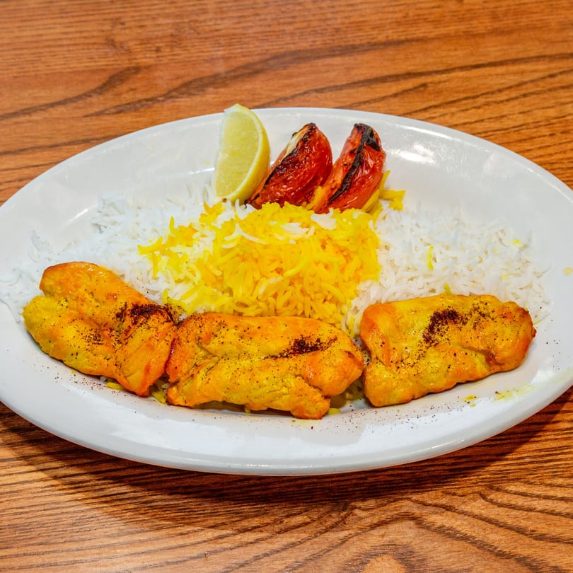 Persian chicken kabob with white and yellow rice, grilled tomatoes, and a lemon wedge.