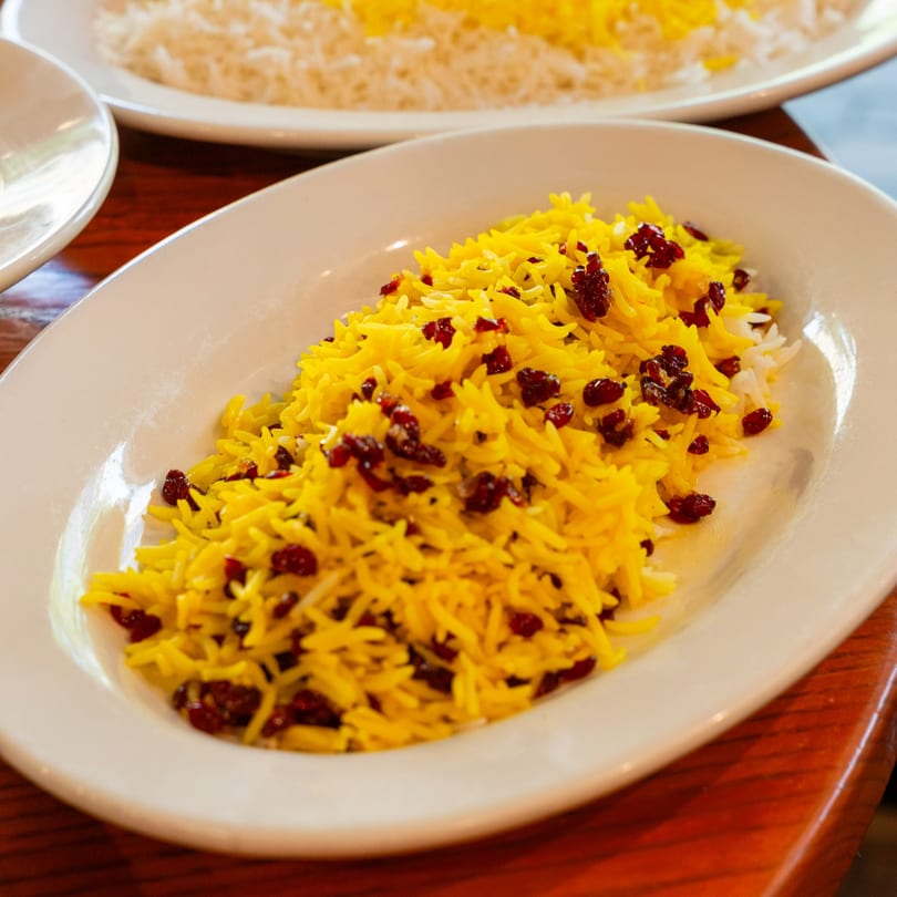A white oval plate filled with yellow Persian rice and red barberries on a wooden table.