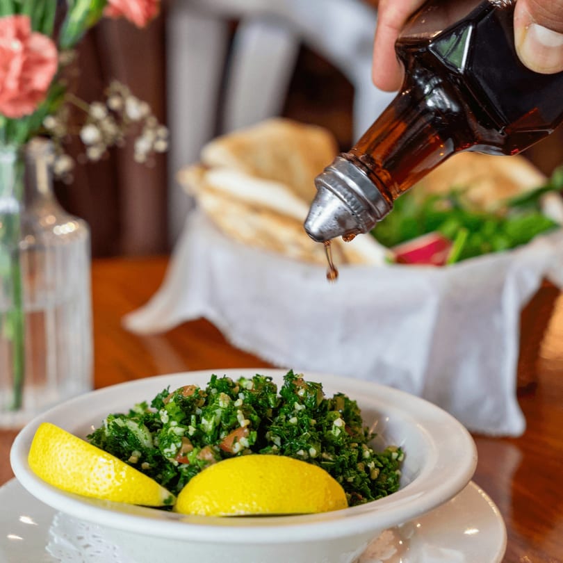 A hand drizzles dressing over a fresh Persian tabbouleh salad with lemon wedges.