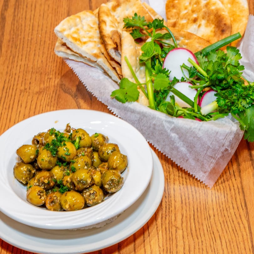 A white bowl of green olives with herbs and a basket of Persian flatbread with fresh herbs and radishes.
