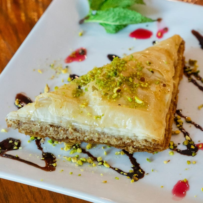 A triangular slice of baklava, topped with chopped pistachios, on a white plate with chocolate and berry drizzles.