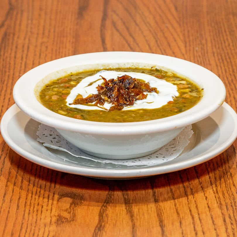 A bowl of Persian Ash Reshteh soup, topped with white cream and crispy fried onions, on a wooden table.
